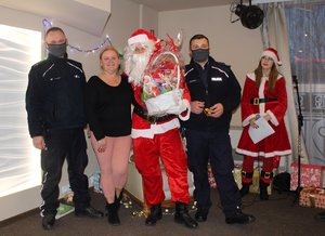 Na zdjęciu widoczny Mikołaj, pomocnica Mikołaja, dwaj umundurowani policjanci stojący na scenie wśród prezentów i kobieta odbierająca kosz pełen słodyczy od Mikołaja.