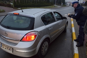 Z prawej - policjant stojący na chodniku przekazuje kierowcy maskotkę przez otwarte okno w samochodzie. Po lewej na pierwszym planie srebrny samochód.