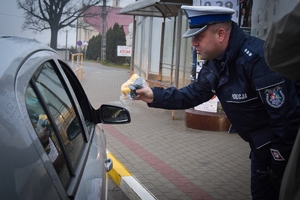 Policjant stojący na chodniku przekazuje kierowcy maskotkę przez otwarte okno w samochodzie.