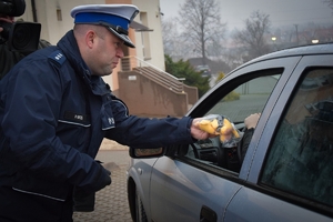 Policjant stojący na chodniku przekazuje kierowcy maskotkę przez otwarte okno w samochodzie.