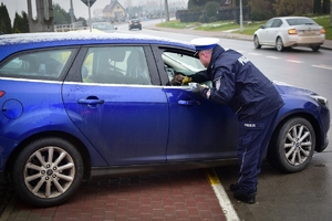 Policjant stojący na chodniku przekazuje kierowcy maskotkę przez otwarte okno w samochodzie.