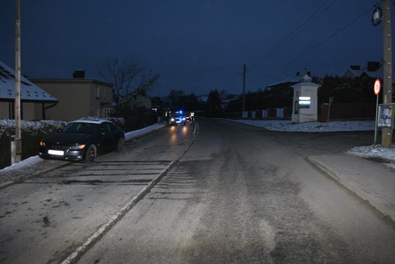Zdjęcie wykonane wieczorem. Po lewej stronie na pierwszym planie samochód bmw stojący na  poboczu drogi. W tle radiowóz.