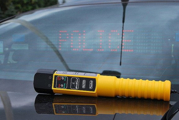 Zjecie kolorowe przedstawia urządzenie do pomiaru alkoholu w wydychanym powietrzu które leży na tylnej klapie samochodu policyjnego nieoznakowanego z włączonym na szybie napisem POLICJA w Czerwonym kolorze. Urządzenie jest koloru żółtego z czarnym ustnikiem ,