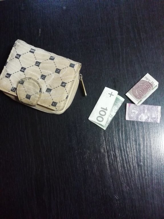 Zdjęcie kolorowe przedstawia blat stołu koloru czarnego na którym po lewej stronie położony jest portfel damski w białym kolorze z jasno brązowymi kwadratami , a po lewej widoczne są złożone banknoty w nominale 100, 20, i 10 zł.