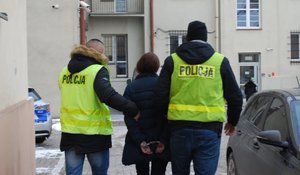 Na zdjęciu dwaj nieumundurowania policjanci w żółtych kamizelkach z napisem POLICJA na plecach prowadzą przez dziedziniec do radiowozu podejrzana kobietę.