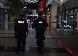 Na zdjęciu umundurowania policjanci idą holem galerii handlowej. Na plecach napis policja. W tle witryny sklepów