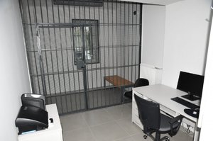 pomieszczenie przejściowe z miejscem dla osób zatrzymanych, oddzielone kratą, po lewej na stoliku urządzenia: alkotest i narkotest, po prawej komputer i drukarka