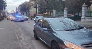 Zdjęcie kolorowe wykonane w porze dziennej przedstawia miejsce zdarzenia drogowego z udziałem pieszej. Zdjęcie przedstawia ul. Krasińskiego w Przemyślu , na przednim planie widoczny jest pojazd m-ki peugeot w kolorze popielatym  a za nim pojazd policyjny z właczonymi światłami błyskowymi.