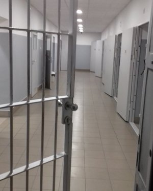 Zdjęcie kolorowe przedstawia pomieszczenie dla zatrzymanych w KMP w Przemyślu . Po prawej stronie zdjęcia widoczne są pootwierane drzwi do pomieszczeń dla zatrzymanych a po lewej stronie widoczne drzwi do urządzeń sanitarnych. Na przednim planie fotografii widoczne są kraty.  Pomieszczenie jest w kolorze popielatym.