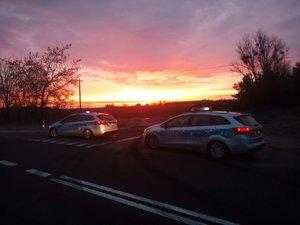 Dwa policyjne radiowozy na tle zachodzącego słońca. Pojazdy stoją na skrzyżowaniu dróg.