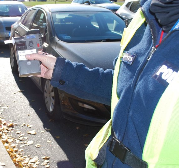 Na zdjęciu umundurowany policjant ruchu drogowego trzyma w wyciągniętej dłoni urządzenie do kontroli trzeźwości. W tle zaparkowane przy krawężniku jezdni samochody.