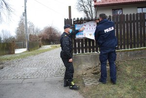 Zdjęcie kolorowe wykonane w porze dziennej przedstawia wjazd do świetlicy wiejskiej w miejscowości Kuńkowce. Przed wjazdem po prawej r widoczny jest policjant umundurowany oraz strażak, mężczyźni wieszają plakat na tablicy ogłoszeń –w tle widoczny budynek świetlicy.