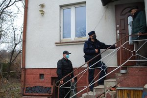 Zdjęcie kolorowe wykonane w porze dziennej-  przedstawia wejście do domu mieszkalnego. Przy drzwiach starszy mężczyzna ubrany w ciemną kurtkę oraz brązowe spodnie, na głowie ma czapkę w kolorze czarnym. Na schodach przed starszym mężczyzną stoi policjant umundurowany oraz strażak.