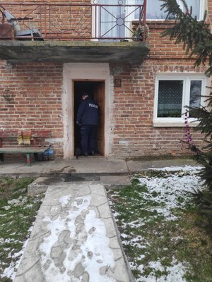 Zdjęcie kolorowe wykonane w porze dziennej-  przedstawia wejście do domu mieszkalnego. W drzwiach widoczny policjant wchodzący do domu umundurowany. Dom jest budowany z ciegieł które są widoczne na zdjęciu