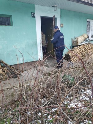 Zdjęcie kolorowe wykonane w porze dziennej-  przedstawia wejście do domu mieszkalnego. W drzwiach widoczna jest starsza kobieta ubrana w ciemnoniebieska bluzkę oraz bezrękawnik w kolorze jasnym przed kobietą stoi policjant umundurowany. Dom jest budowany w kolorze zielonym.