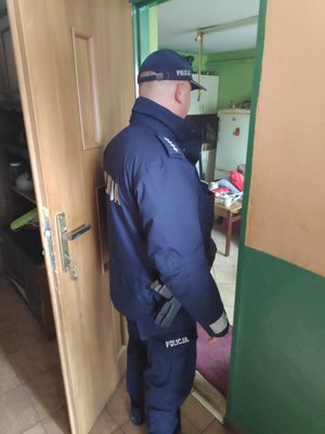 Zdjęcie kolorowe wykonane w porze dziennej przedstawia policjanta umundurowanego , który stoi w drzwiach mieszkania