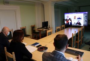 w sali, przy stole widoczne od tyłu sylwetki trzech osób, w oddali ekran, na którym widać sylwetki kilku osób biorących udział w wideokonferencji