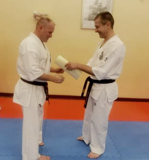 Na zdjęciu znajduje się dwóch mężczyzn ubranych w tradycyjny strój karateki czyli kimono, z których jeden wręcza drugiemu certyfikat na sali treningowej
