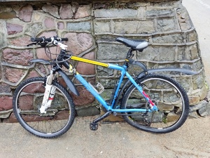 Zdjęcie przedstawia rower w kolorze niebieskim z żółtymi i białymi wstawkami. Pojazd uczestniczył w zdarzeniu drogowym.