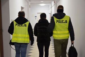 Dwóch funkcjonariuszy, w kamizelkach z napisem POLICJA, prowadzi przez korytarz zatrzymaną kobietę