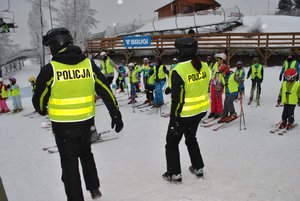 Na pierwszym planie widoczna dwójka funkcjonariuszy stojących na stoku narciarskim, w tle użytkownicy stoku