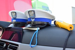 Zdjęcie kolorowe przedstawia podszybie samochodu na którym leżą dwie czapki policjantów ruchu drogowego oraz urządzenie do pomiaru alkoholu w wydychanym powietrzu o nazwie „ alcoblov” . Urządzenie jest koloru żółtego z czarnym ustnikiem