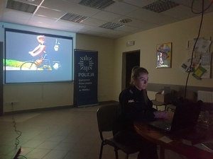 policjantka w czasie zajęć online, siedzi przy komputerze, za policjantką znajduje się ekran multimedialny na którym wyświetlana jest prezentacja