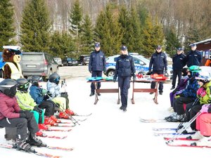 policjantka prowadzi prelekcję, po jej prawej i lewej stronie siedzą dzieci z kombinezonach narciarskich oraz z zapiętymi nartami