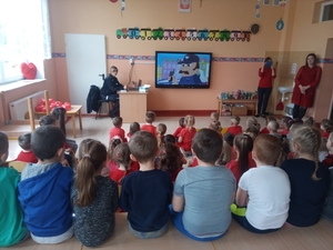 Na zdjęciu znajduje się policjantka, nauczycielka oraz dzieci w szkolnej klasie oglądający prezentację na ekranie