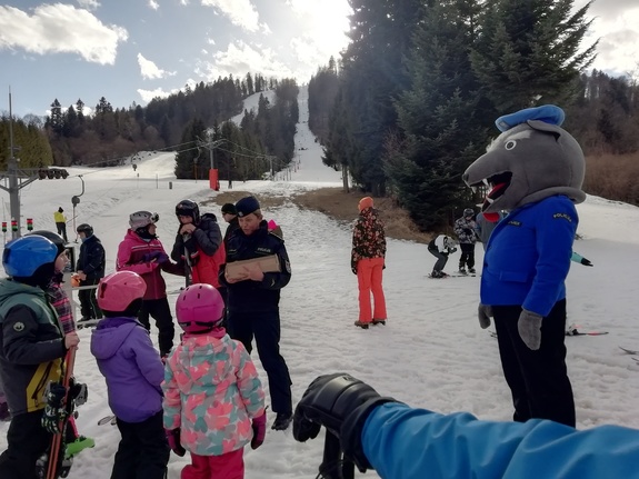 Policjantka (w centrum kadru) rozmawia z dziećmi na stoku narciarskim. Po prawej policyjna maskotka - wilk w policyjnym mundurze. W tle widać stok i wyciąg.