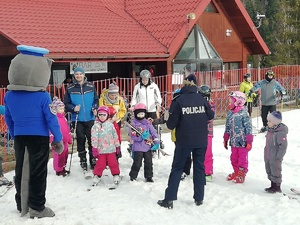 Policjantka (tyłem w centrum kadru) rozmawia z dziećmi na stoku narciarskim.