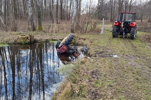 Zdjęcie wykonane w ciągu dnia. W środku kadru ciągnik leżący kołami do góry zanurzony do połowy w stawie (z wody wystają koła i część błotnika). Po prawej stronie drugi ciągnik, czerwony, który za pomocą liny wydobywa przewrócony pojazd ze zbiornika wodnego. W tle drzewa.