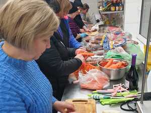 Kobiety przygotowujące posiłek w kuchni. Stoją po lewej stronie, przy stole, na którym są produkty spożywcze. Kobiety obierają warzywa, wędliny.