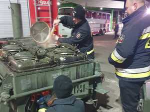 Strażacy rozpalają ogień w kuchni polowej i mieszają gotującą się grochówkę. Jeden ze strażaków otwiera kotły. W tle wóz strażacki i autobus.