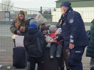 Funkcjonariusz podaje dzieciom siedzącym na walizkach policyjną maskotkę - misia policjanta.