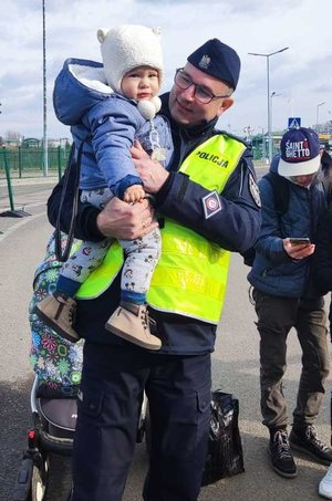 Zdjęcie kolorowe przedstawia policjanta który pomaga uchodźcom z Ukrainy  w przenoszeniu bagaży