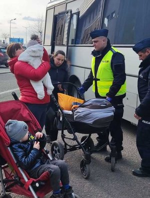 Zdjęcie kolorowe przedstawia policjanta który pomaga uchodźcom z Ukrainy  w przenoszeniu bagaży