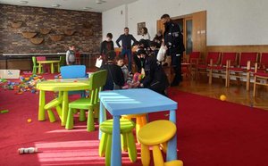 Zdjęcie wykonane w sali zabaw. Na pierwszym planie kolorowe stoliki i krzesełka. Policjanci przekazują zabawki dzieciom.