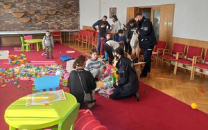 Policjantka (bokiem) przekazuje piłkę małemu chłopcu. W tle na podłodze rozsypane klocki, wśród dzieci stoją także opiekunowie.