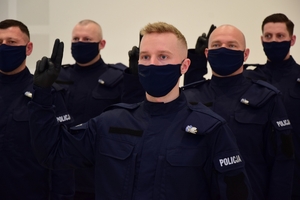 Nowo przyjęci policjanci podczas ślubowania, z podniesioną prawą ręką, stoją w maseczkach.