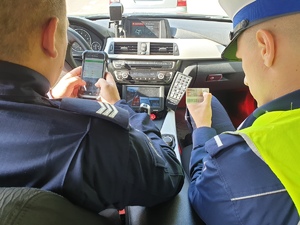 zdjęcie poglądowe, dwóch policjantów w radiowozie podczas kontroli drogowej, jeden z nich trzyma w rękach prawo jazdy kierującego poddanego kontroli