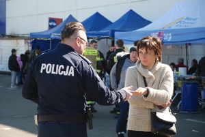 Zdjęcie kolorowe wykonane w porze dziennej przedstawia policjanta który rozdaje ulotki