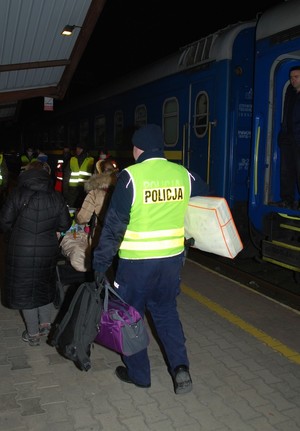 Zdjęcie kolorowe wykonane w porze nocnej przedstawia dworzec PKP w Przemyślu- na zdjęciu widoczny policjant umundurowany który pomaga uchodźcom w przenoszeniu bagaży