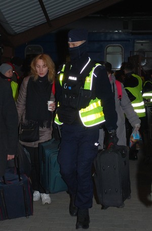 Zdjęcie kolorowe wykonane w porze nocnej przedstawia dworzec PKP w Przemyślu- na zdjęciu widoczny policjant umundurowany który pomaga uchodźcom w przenoszeniu bagaży