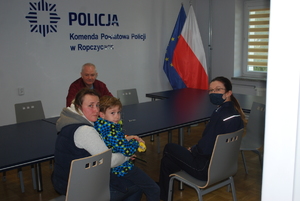 Od lewej na pierwszym planie siedzi kobieta, trzymająca dziecko na kolanach, po jej prawej stronie siedzi policjantka, natomiast po ich przeciwnej stronie siedzi mężczyzna