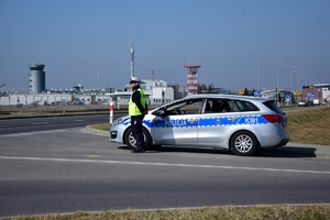 Policjant ruchu drogowego stoi przy radiowozie (po prawej stronie) przy wyjeździe z bocznej ulicy. W tle budynki i infrastruktura lotniska.