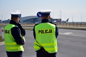 Po lewej trzej policjanci ruchu drogowego stojący przy drodze. W tle zabudowania lotniska.