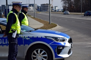 Policjanci ruchu drogowego stoją przy radiowozie (po lewej stronie) przy wyjeździe z bocznej ulicy. W tle ulica i zabudowania lotniska.