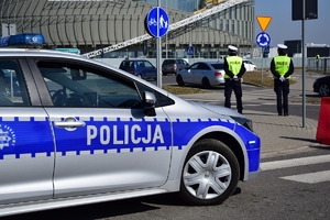 Na pierwszym planie po lewej policyjny radiowóz. W tle po prawej dwoje policjantów ruchu drogowego stojący obok przejścia dla pieszych.