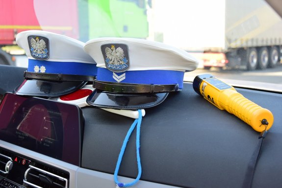 Dwie policyjne czapki funkcjonariuszy ruchu drogowego oraz urządzenie służące do sprawdzania stanu trzeźwości, położone na desce rozdzielczej radiowozu